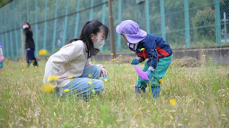 春の野原で学生とこどもがむしを捕まえようとしている。